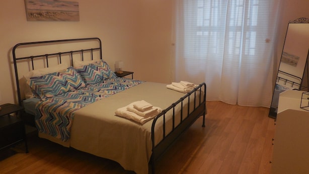 A bedroom with a bed, a nightstand, a lamp, a window with curtains, and a painting on the wall. - Vrbo Property (Pozzuoli)