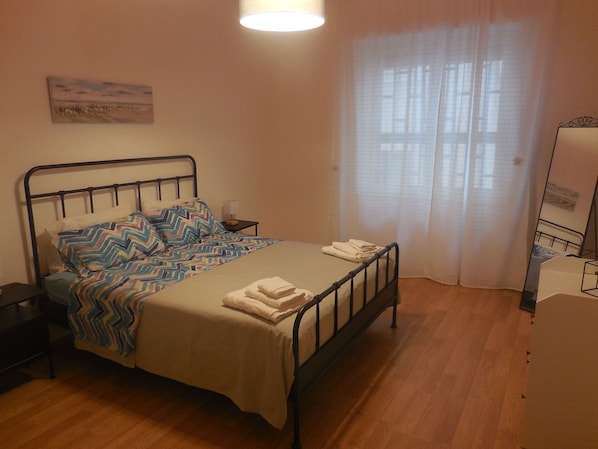 Ein Schlafzimmer mit einem Bett, einem Nachttisch, einer Lampe, einem Fenster mit Vorhängen und einem Bild an der Wand.