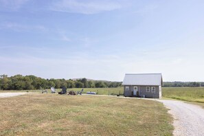 A small house with a metal roof, a gravel driveway, and a grassy field with a few chairs and a table. - Enjoy this peaceful 1-bedroom cabin in the country for a relaxing stay. (Lamar)