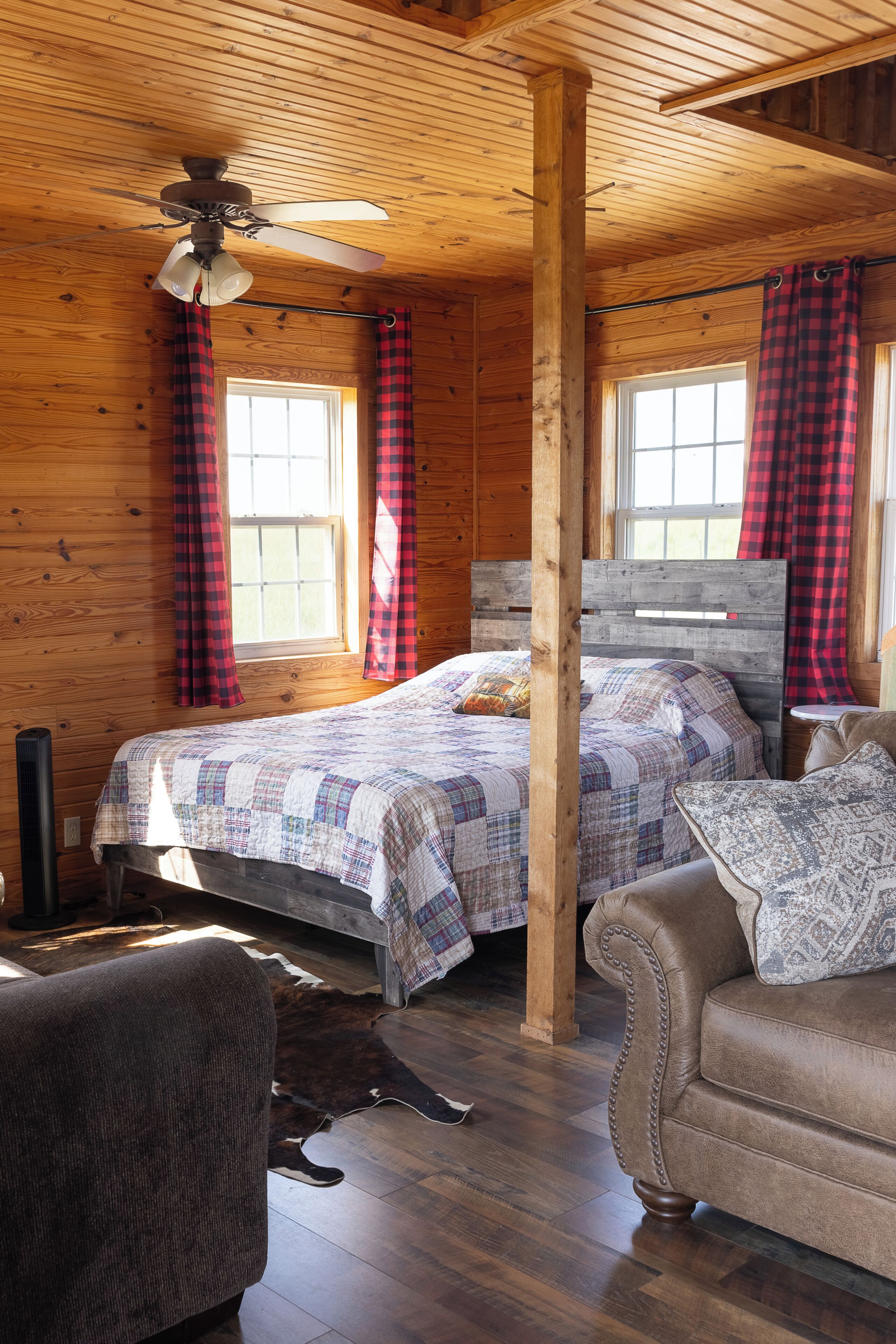 A wooden cabin room with a bed, two windows with red and black checkered curtains, a ceiling fan, and a wooden floor.