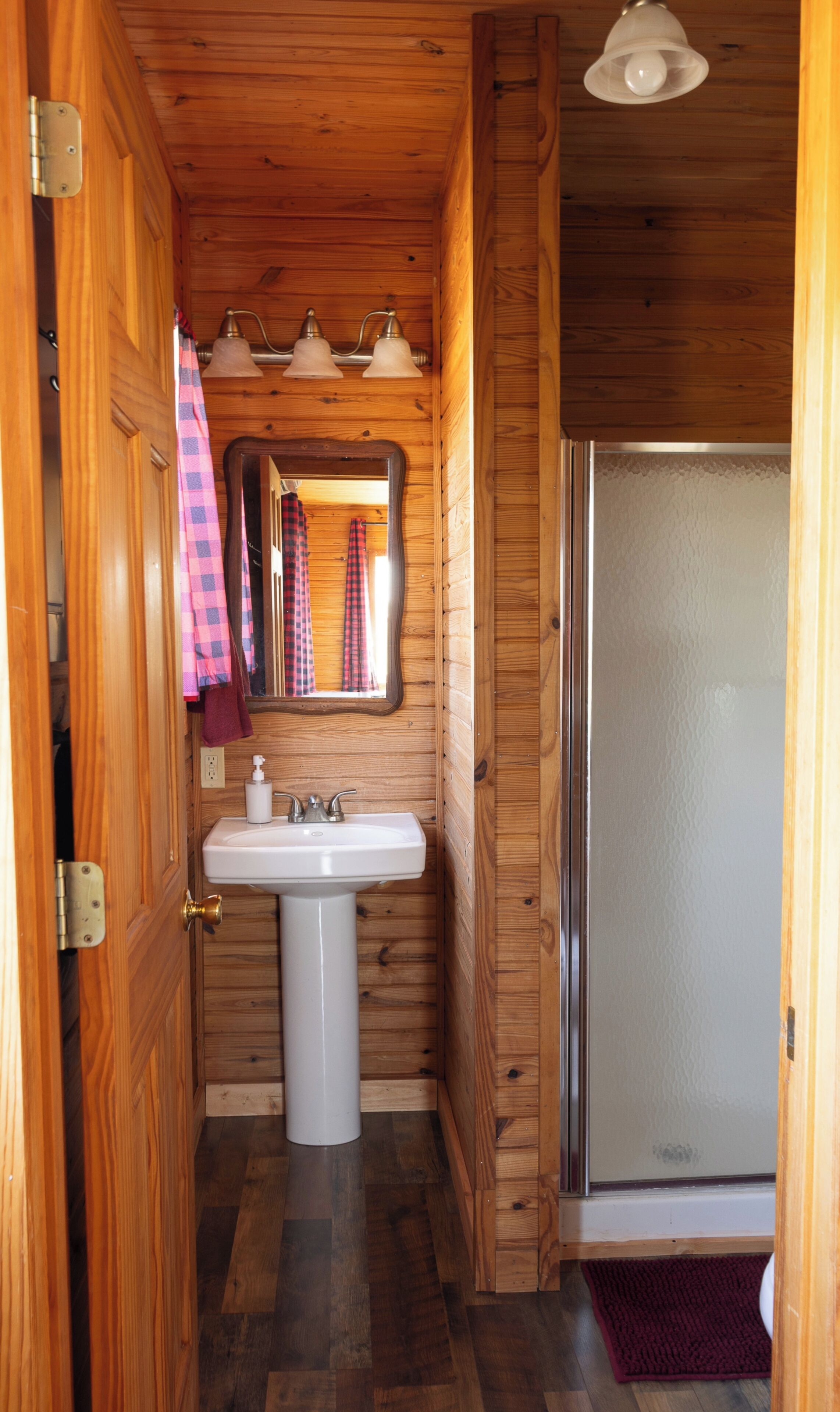 Un interior de madera con un lavabo blanco, un espejo y una puerta con una toalla colgando.