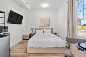 A hotel room with a bed, a TV mounted on the wall, a bedside table with a lamp, and a view of the outside through a window. - Downtown Edmonton Studio | AC, Parking (Edmonton)