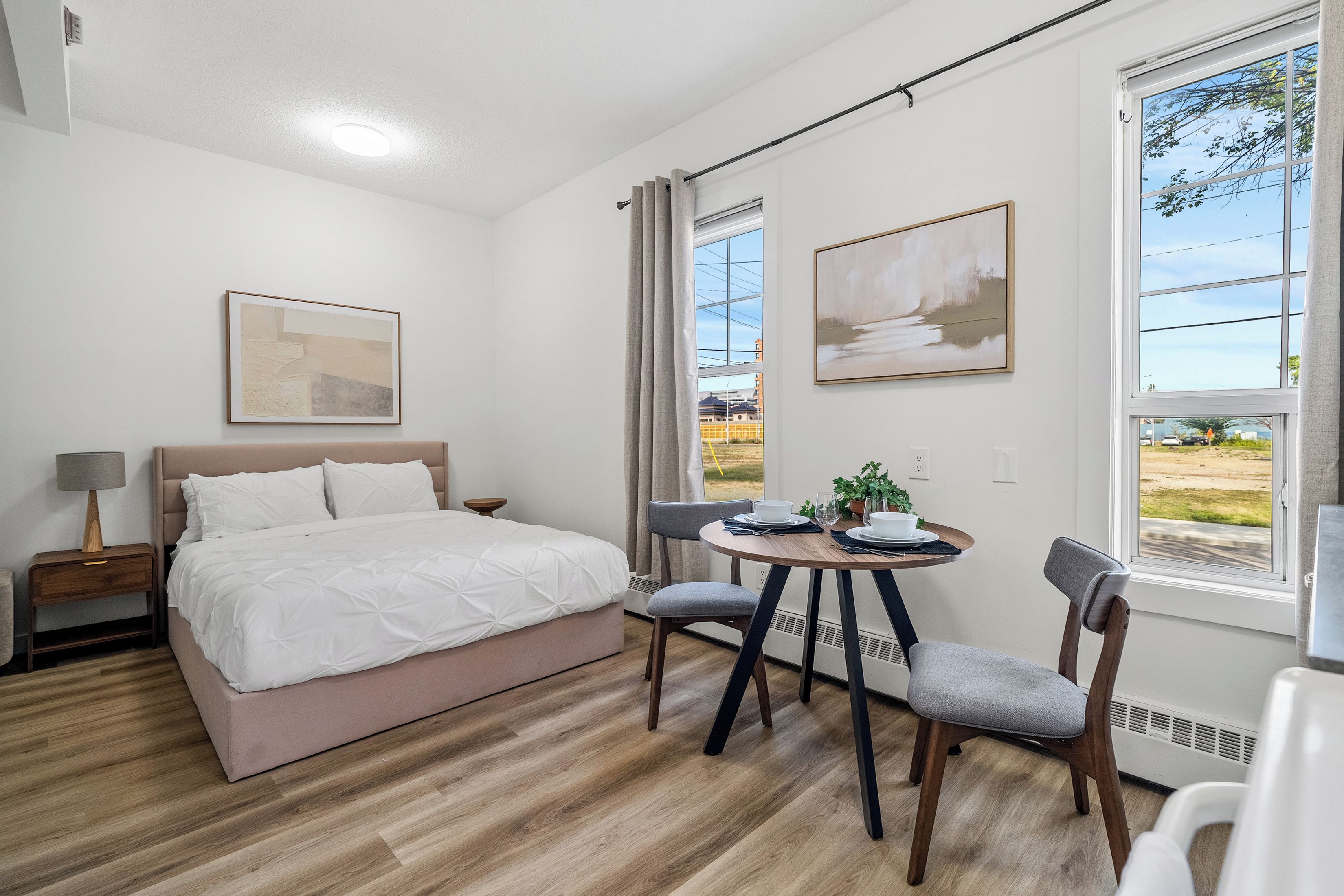 A hotel room with a bed, a bedside table with a lamp, a dining table with chairs, and two windows with curtains.
