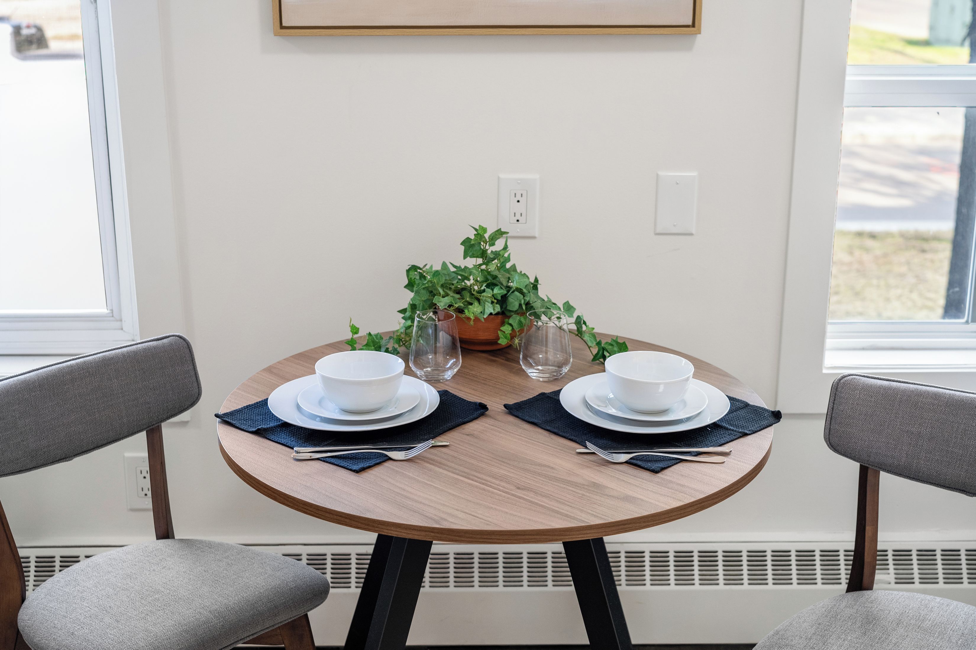 Una mesa de comedor con dos cuencos blancos, dos platos y dos tenedores, sobre individuales azul oscuro. Hay una planta en maceta con hojas verdes en la mesa.