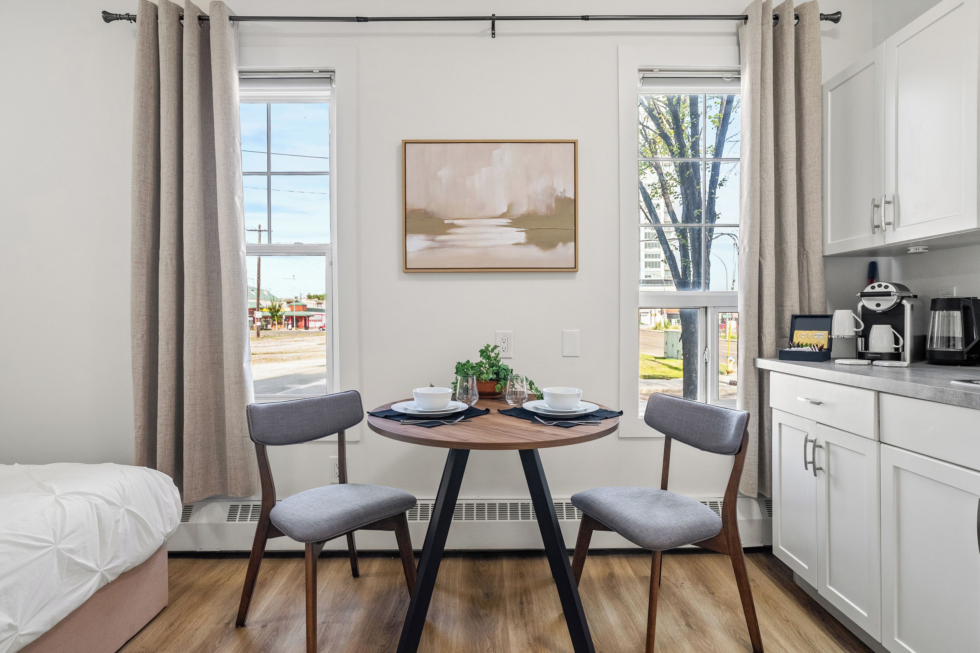 A modern kitchen with a dining area, featuring a table with chairs, a painting on the wall, and a view of the outside.