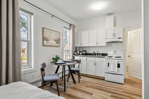 A compact kitchen with white cabinets, a dining table with chairs, and a small window. - Downtown Edmonton Studio | AC, Parking (Edmonton)