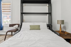 A bunk bed room with a white bedspread, a green pillow, a wooden nightstand with a lamp, and a chair with a white cushion. - Studio w Queen Murphy Bed | Parking, Wifi, Dining (Edmonton)
