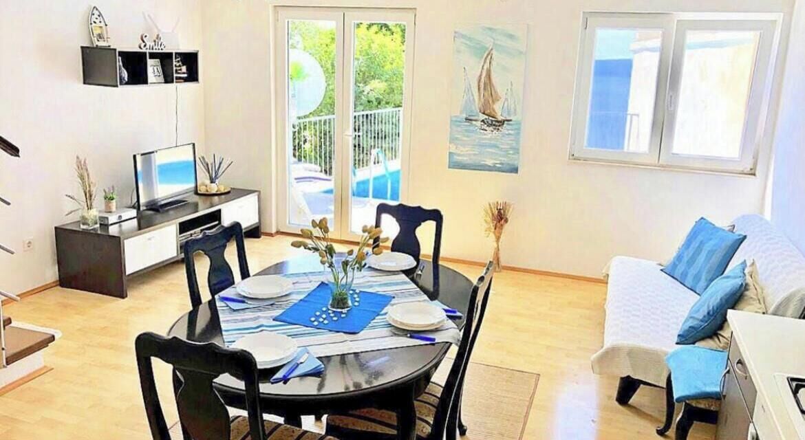 A dining area with a round table, chairs, and a TV on a stand. There is a balcony with a view of greenery and a sailboat painting on the wall.
