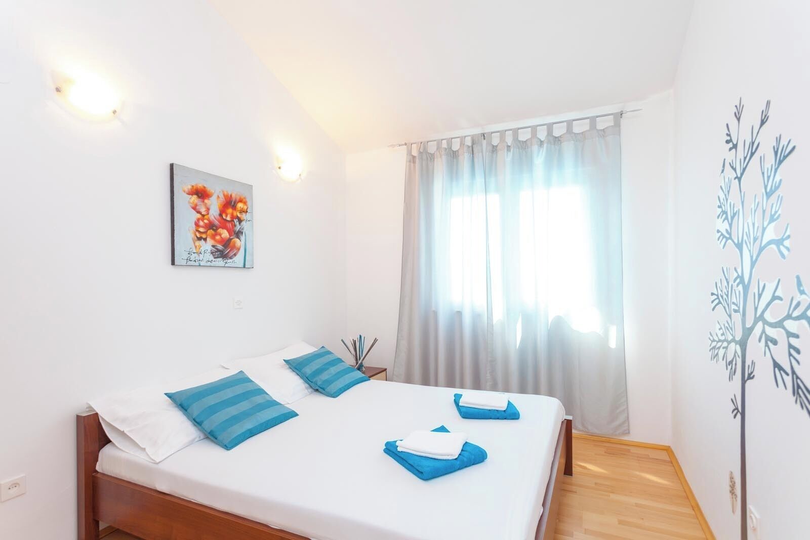 A bedroom with a bed, two pillows, a painting, a window with curtains, and a wall decoration.