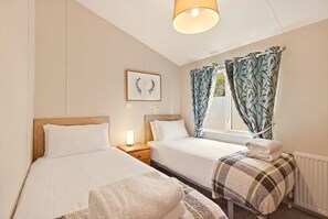 Hotellihuone, jossa on kaksi sänkyä, yöpöytä lampun kanssa, ikkuna verhoilla ja patteri.