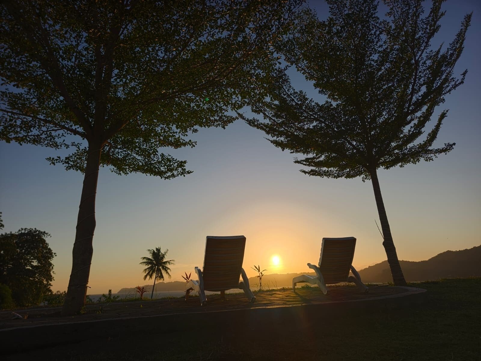 Deux fauteuils de jardin sont placés face à un coucher de soleil, avec un palmier et une montagne en arrière-plan.