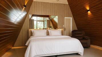 A bedroom with a sloped ceiling, a bed with white bedding, a brown leather chair, and a window with curtains.