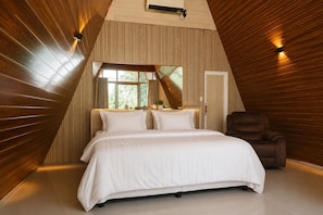 A bedroom with a sloped ceiling, a bed with white bedding, a brown leather chair, and a window with curtains.