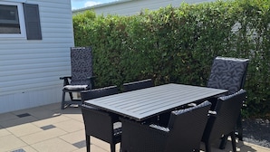 En udendørs patio med et bord og stole, et mindre overdækket siddeområde og en hæk i baggrunden.