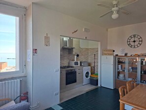 A compact kitchen with a washing machine, a microwave, and a stove. There is a dining area with a table and chairs. A clock is visible on the wall. - Vrbo Property (Lido di Pomposa)