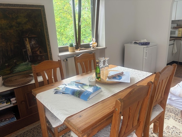 Un comedor con una mesa de madera, sillas, un cuadro en la pared, un ventanal con vista a árboles y una zona de cocina al fondo.