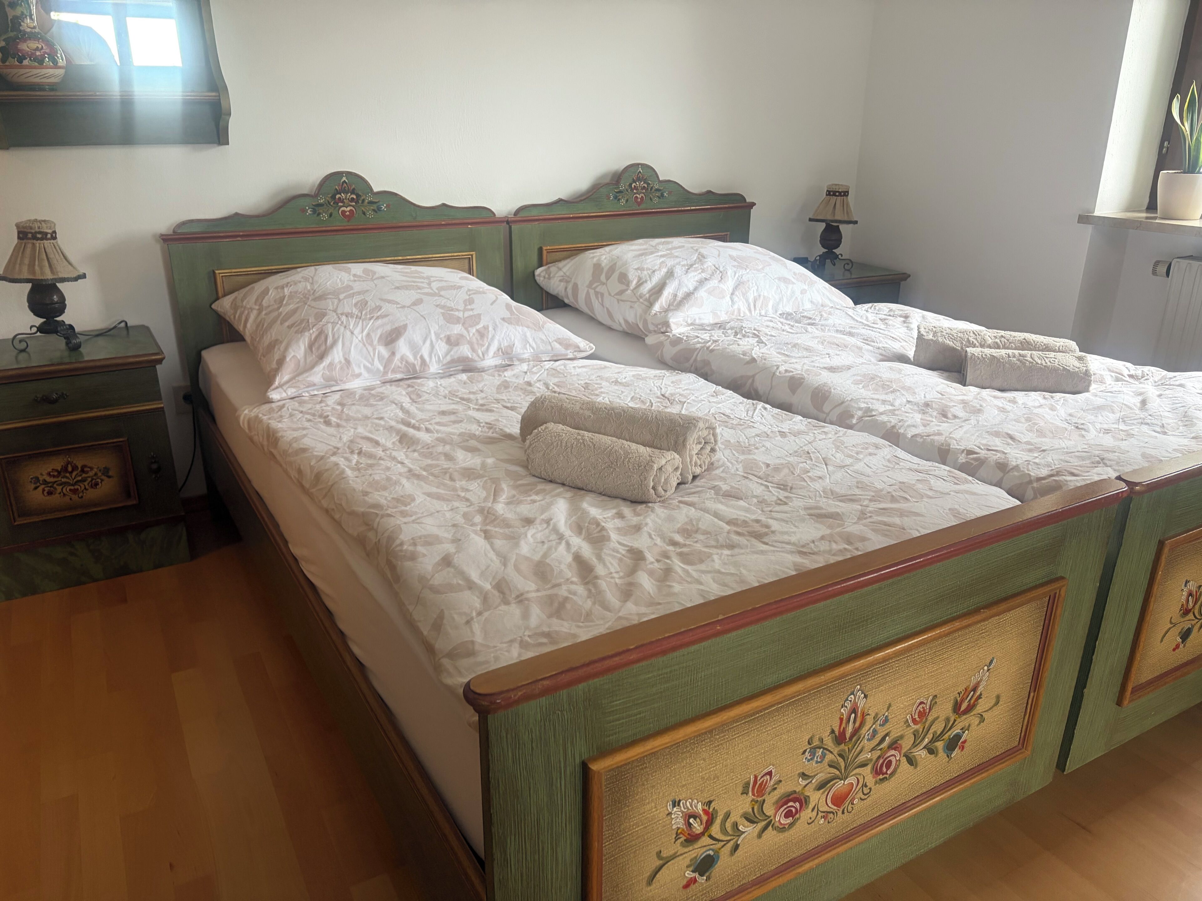 Ein Doppelbett mit floralem Kopfteil und Fußteil, zwei Nachttische mit Lampen und ein Fenster mit Vorhängen.