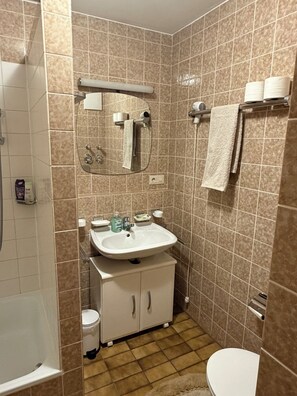 Un baño con lavamanos blanco, espejo e inodoro. Hay un perchero con toallas y un cabezal de ducha.