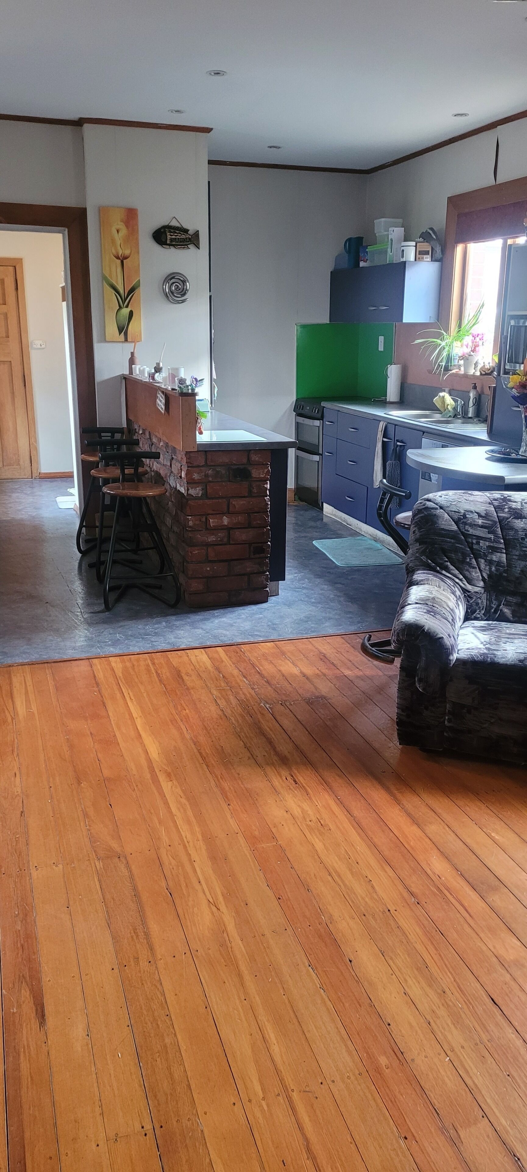 A living room with a wooden floor, a brick fireplace, a green kitchen area, and a dining area with stools.