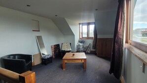 A room with a sloped ceiling, a wooden table, a black chair, a window with curtains, and a view of a residential area.