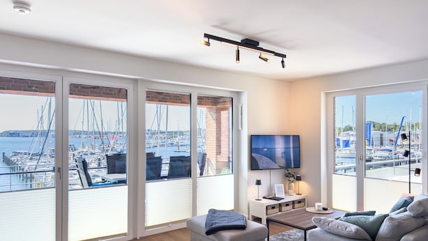 A modern living room with a view of a marina, featuring a sofa, a coffee table, and a flat-screen TV. - Listing No. 303 “Baltique” in Travemünde, featuring a large south-facing loggia and harbor views (Travemünde)
