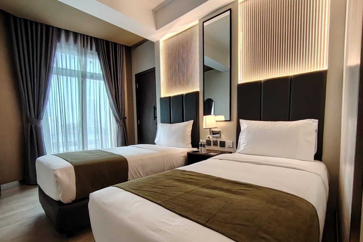 Ein Hotelzimmer mit zwei Betten, einem großen Spiegel, einem Nachttisch und einem Fenster mit Vorhängen.