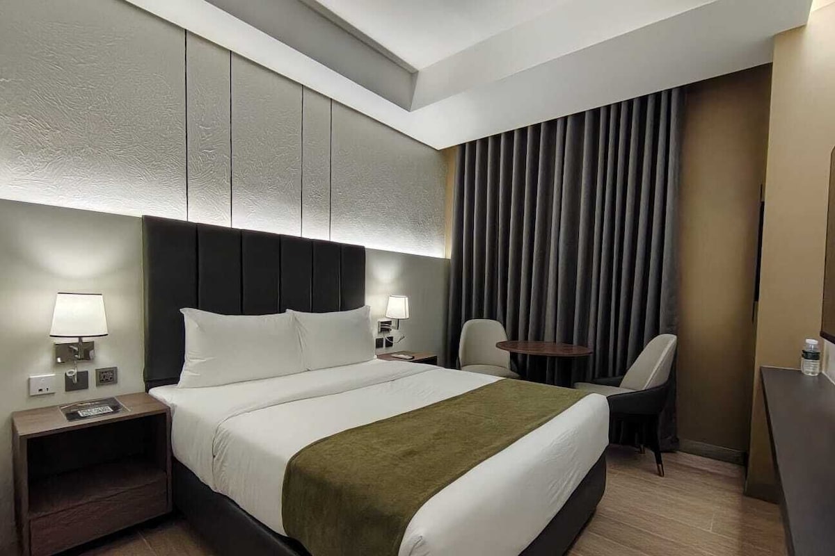 Ein Hotelzimmer mit Bett, Nachttischen, einem Schreibtisch, einem Sessel und einem großen Fenster mit Vorhängen.