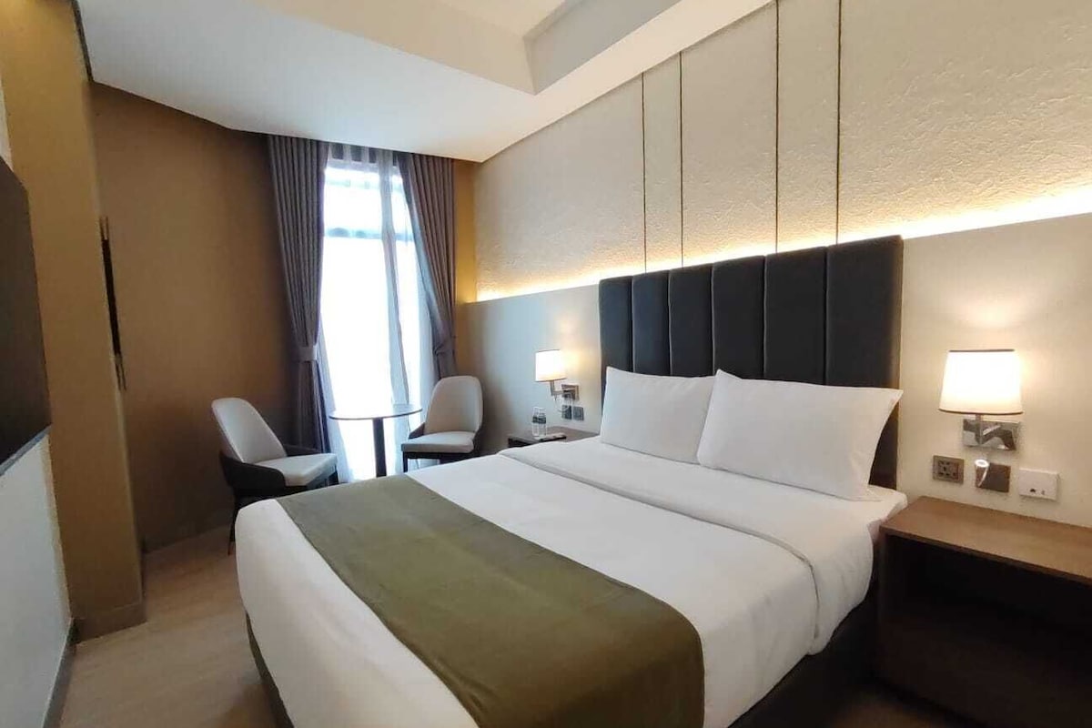Ein Hotelzimmer mit einem großen Bett, Nachttischen, einem Sessel und einem Fenster mit Vorhängen.