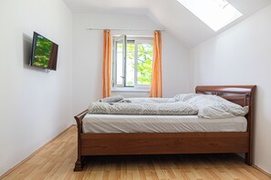 A bedroom with a wooden bed, a TV mounted on the wall, a window with orange curtains, and a skylight.