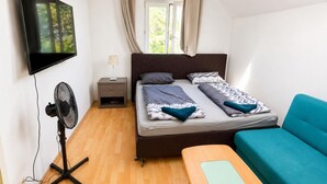 A bedroom with a bed, a TV, a fan, a window with curtains, and a blue sofa. - Private Villa Room for 4 | Garden Access (Vienna)