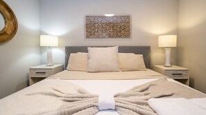 Cama bem arrumada com um cobertor grande dobrado ao centro, acompanhada por duas luminárias de mesa, uma cabeceira e um elemento decorativo na parede acima da cama.