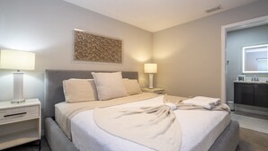 Habitación de hotel con cama, mesitas de noche, una obra de arte colgada en la pared, un baño con espejo y lavamanos.