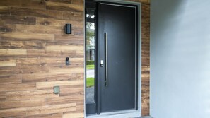 Uma porta moderna com moldura preta, puxador vertical e um pequeno objeto retangular na parede à esquerda.