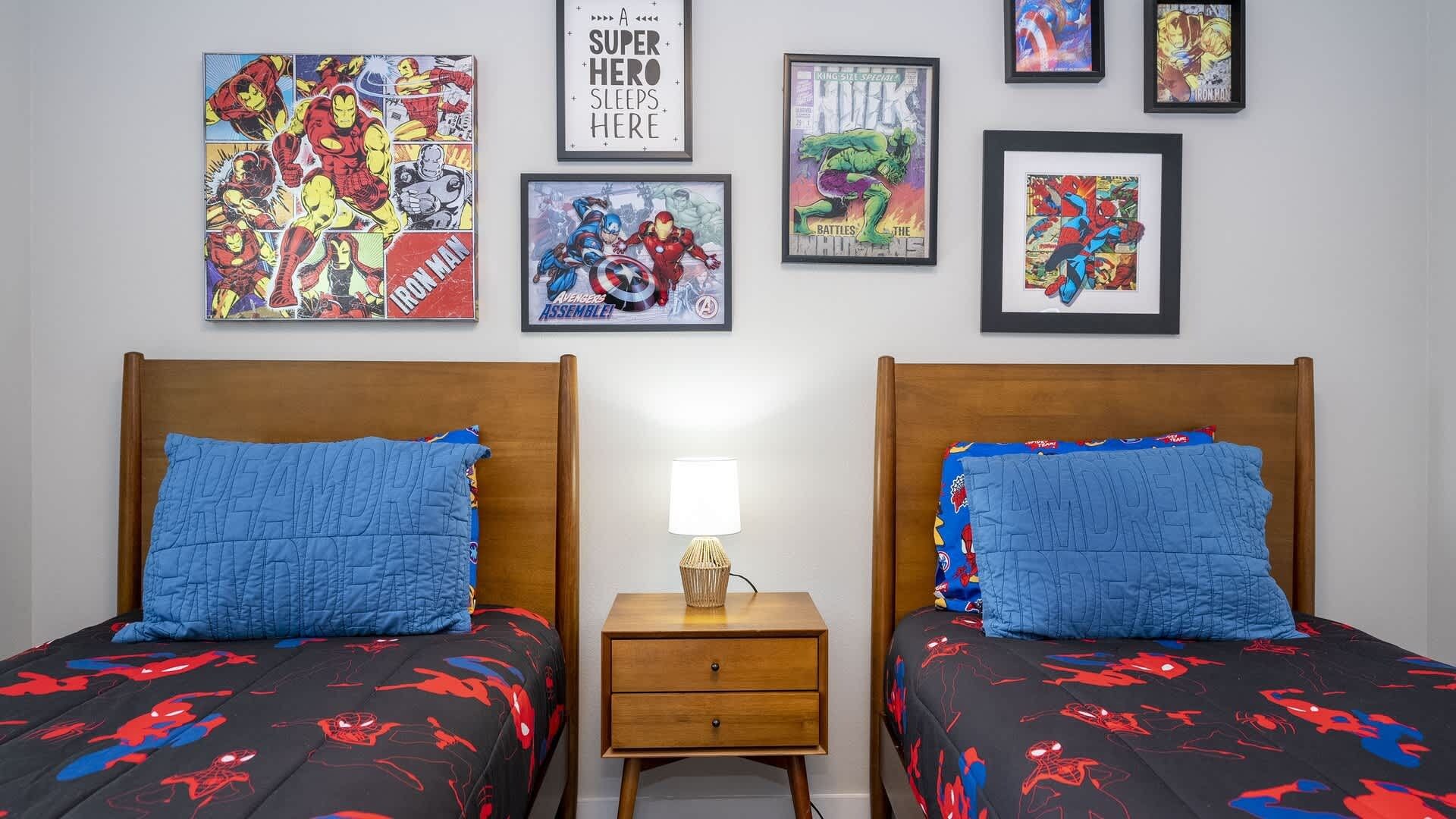 A bedroom with two beds, each with a blue quilted pillow and a Spider-Man themed bedspread. There are framed comic book prints on the wall and a small bedside table with a lamp between the beds.