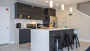 A modern kitchen with dark cabinets, a central island with stools, a coffee maker, and a stainless steel refrigerator. - 4BR Le Reve Pool & Themed Rooms Near Disney (Kissimmee)