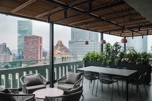 Zona de estar al aire libre con mobiliario de mimbre y una mesa, con vistas a un paisaje urbano de edificios modernos.