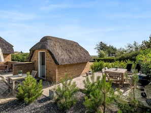 Une charmante maison de campagne au toit de chaume, avec une façade en briques, une terrasse dotée de mobilier d’extérieur et un barbecue.