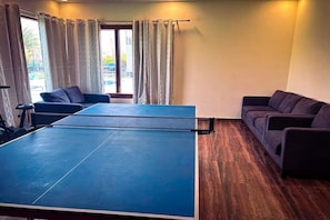 A room with a ping pong table, two sofas, and a window with curtains.