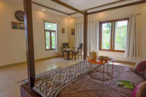 Quarto com cama, mesa com chaleira e cadeira. Janelas com cortinas.