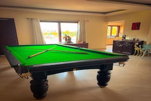 A pool table in a room with a view through a window, a cabinet, and a door.