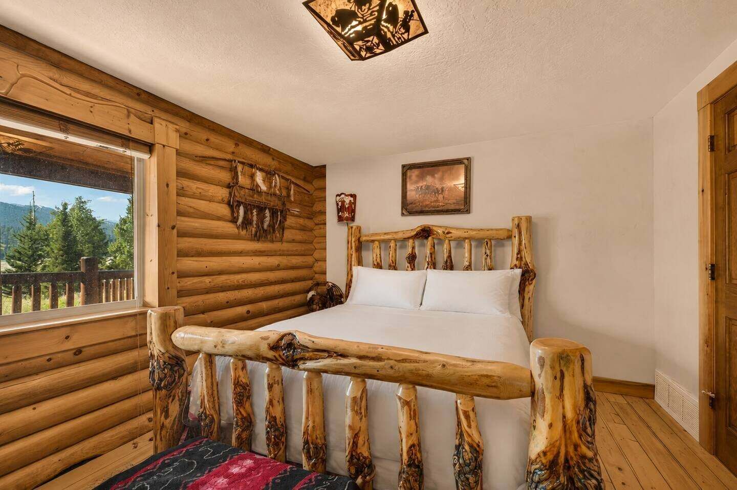 A log cabin room with a bed, a window with a view of trees, and a wooden headboard.