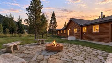 A log cabin with a stone fire pit and benches, surrounded by a grassy yard and trees.