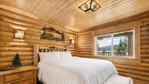 Un dormitorio de cabaña de troncos con cama, mesita de noche, un pequeño árbol de Navidad y ventana con vistas al exterior.