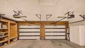A garage with two large white garage doors, overhead lighting, and wooden shelving on the left.