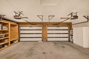 A garage with two large white garage doors, overhead lighting, and wooden shelving on the left.
