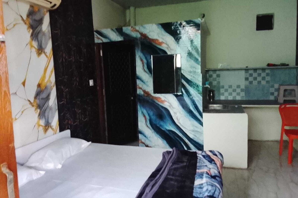 Una habitación con cama, televisión en la pared, una pequeña kitchenette y una silla roja.
