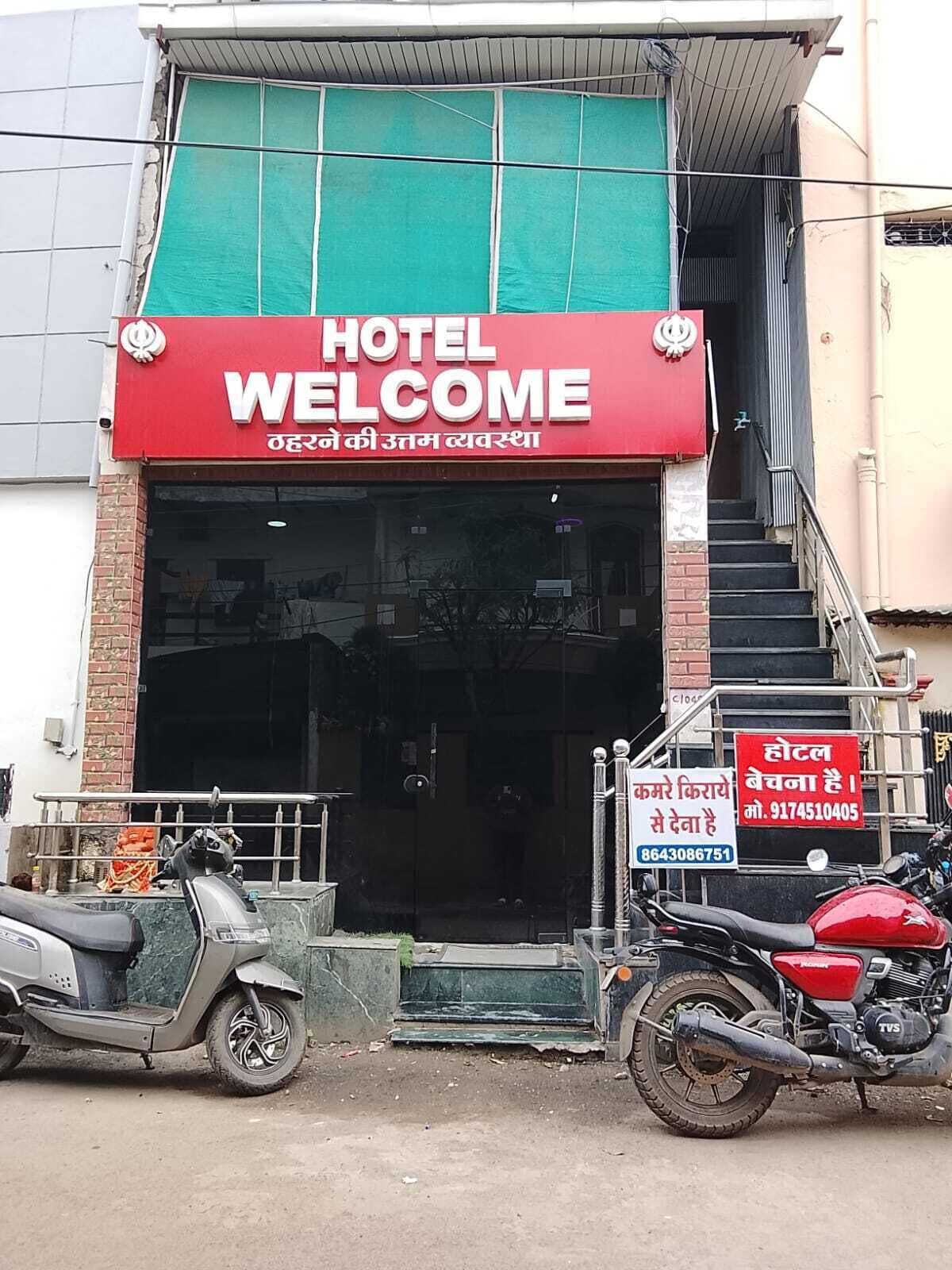 Entrada de un hotel con un letrero rojo que dice 'HOTEL WELCOME' y una placa con información de contacto.