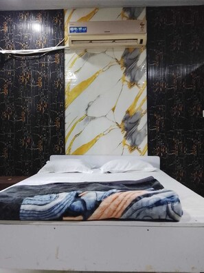 A bedroom with a bed, white pillows, a marble-patterned wall, and an air conditioning unit. - Hotel Welcome (Indore)