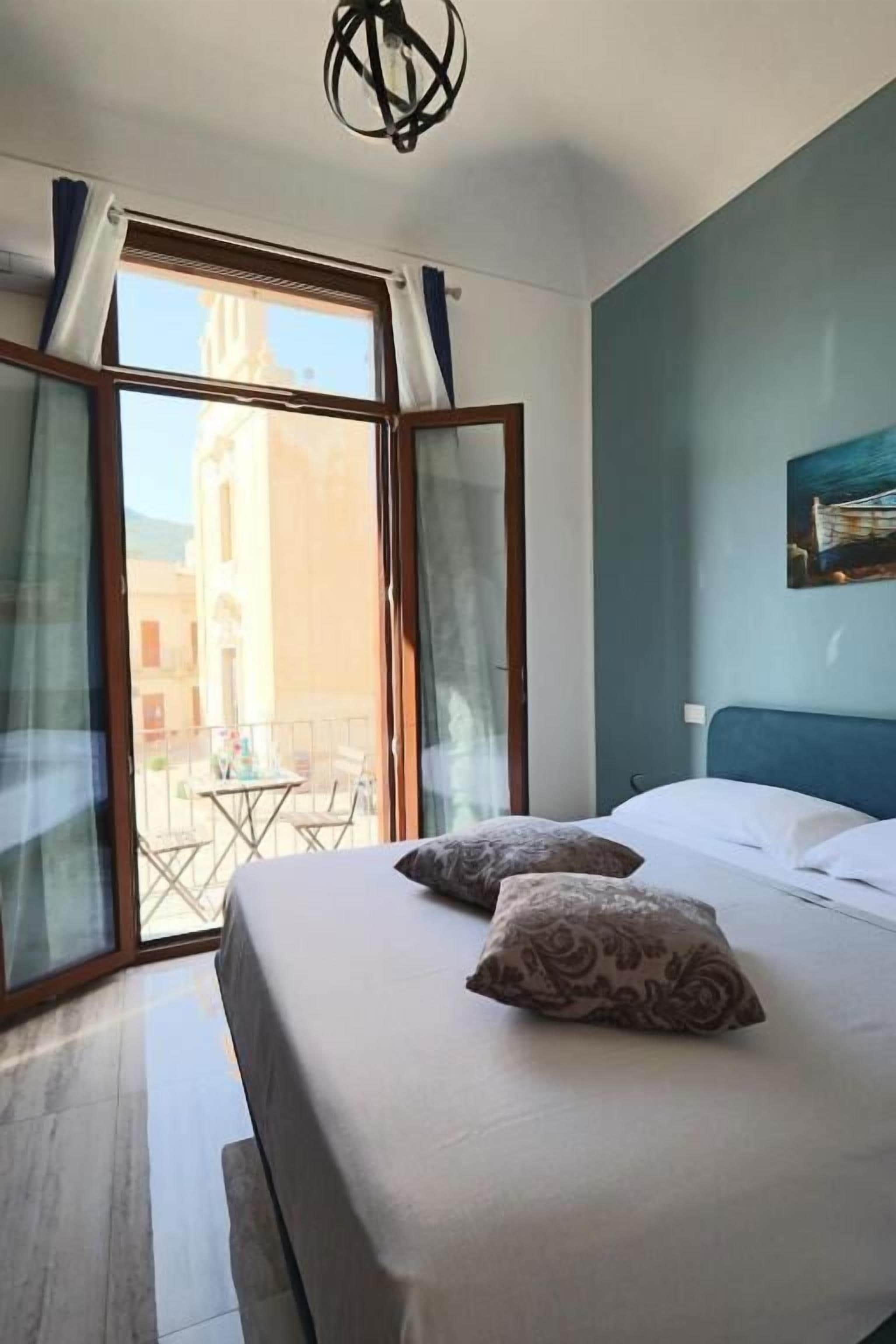 Una camera da letto con un letto, due cuscini, una finestra con tende e vista sugli edifici.
