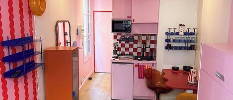 Una cocina compacta con gabinetes rosas, una zona de comedor con una silla amarilla y un ventilador de techo.
